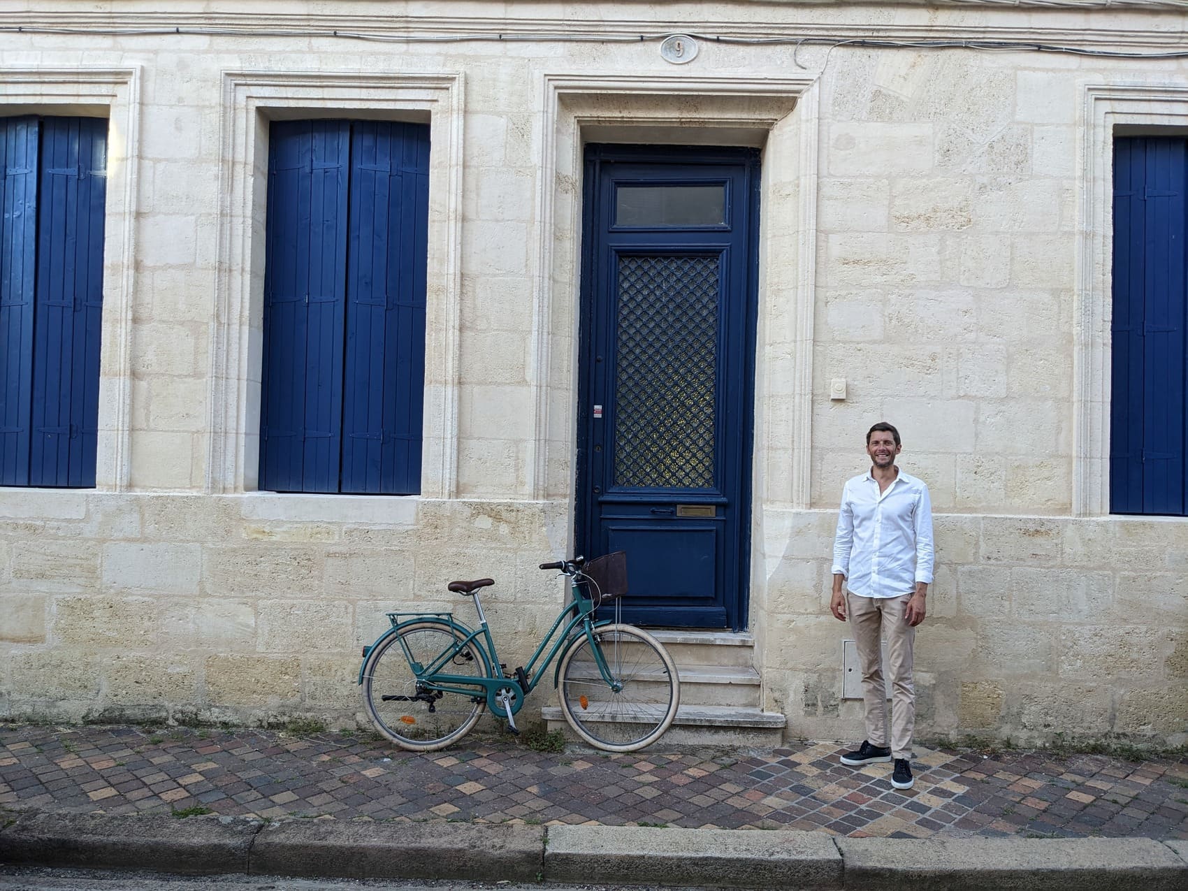 Homme souriant en chemise blanche devant une façade en pierre avec un vélo, illustration de la sérénité apportée par une bonne préparation retraite